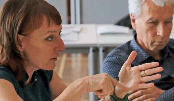 Eine Nahaufnahme von zwei Personen, die in einer Gesprächssituation an einem Tisch sitzen. Im Vordergrund sind Hände zu sehen, die gestikulierend eine Aussage unterstreichen. Auf dem Tisch im Hintergrund liegen mehrere offene Dokumente, und die Umgebung wirkt wie ein Besprechungs- oder Workshopraum.