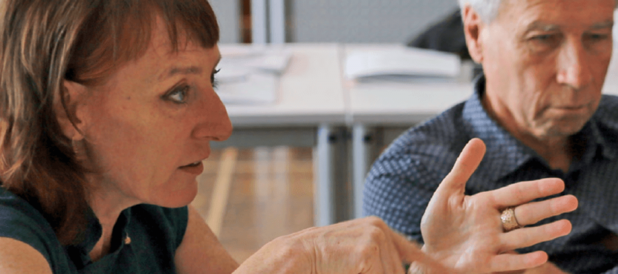 Eine Nahaufnahme von zwei Personen, die in einer Gesprächssituation an einem Tisch sitzen. Im Vordergrund sind Hände zu sehen, die gestikulierend eine Aussage unterstreichen. Auf dem Tisch im Hintergrund liegen mehrere offene Dokumente, und die Umgebung wirkt wie ein Besprechungs- oder Workshopraum.