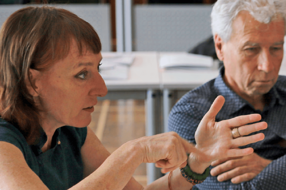 Eine Nahaufnahme von zwei Personen, die in einer Gesprächssituation an einem Tisch sitzen. Im Vordergrund sind Hände zu sehen, die gestikulierend eine Aussage unterstreichen. Auf dem Tisch im Hintergrund liegen mehrere offene Dokumente, und die Umgebung wirkt wie ein Besprechungs- oder Workshopraum.