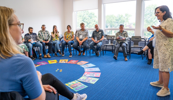 Ein Gruppenraum mit blauem Teppichboden, in dem mehrere Personen in einem Stuhlkreis sitzen. In der Mitte des Kreises liegen bunte Bücher und Karten in einem Halbkreis angeordnet. Eine Person steht am rechten Bildrand und spricht zu der Gruppe, während andere aufmerksam zuhören. Im Hintergrund sind große Fenster mit Blick auf grüne Bäume zu sehen, die viel Tageslicht in den Raum lassen.