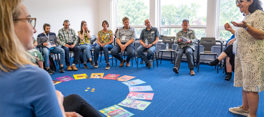 Ein Gruppenraum mit blauem Teppichboden, in dem mehrere Personen in einem Stuhlkreis sitzen. In der Mitte des Kreises liegen bunte Bücher und Karten in einem Halbkreis angeordnet. Eine Person steht am rechten Bildrand und spricht zu der Gruppe, während andere aufmerksam zuhören. Im Hintergrund sind große Fenster mit Blick auf grüne Bäume zu sehen, die viel Tageslicht in den Raum lassen.