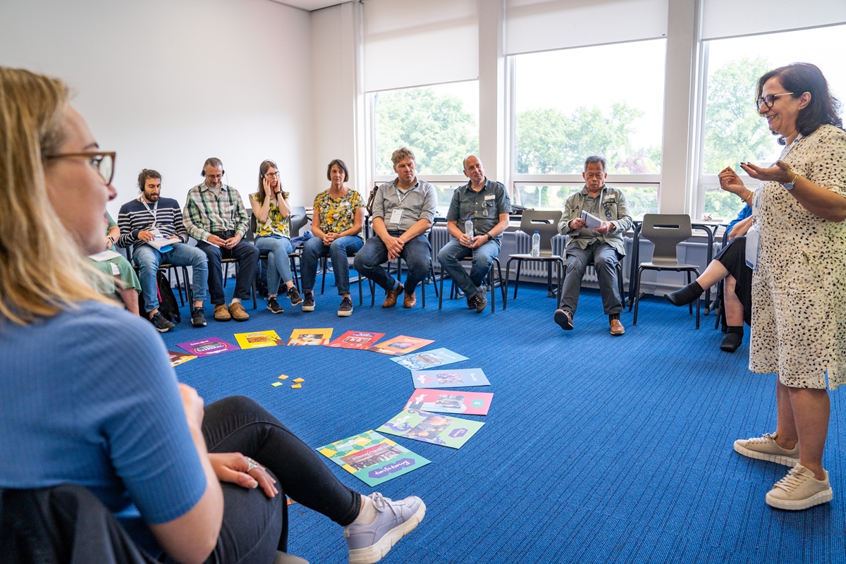 Ein Gruppenraum mit blauem Teppichboden, in dem mehrere Personen in einem Stuhlkreis sitzen. In der Mitte des Kreises liegen bunte Bücher und Karten in einem Halbkreis angeordnet. Eine Person steht am rechten Bildrand und spricht zu der Gruppe, während andere aufmerksam zuhören. Im Hintergrund sind große Fenster mit Blick auf grüne Bäume zu sehen, die viel Tageslicht in den Raum lassen.