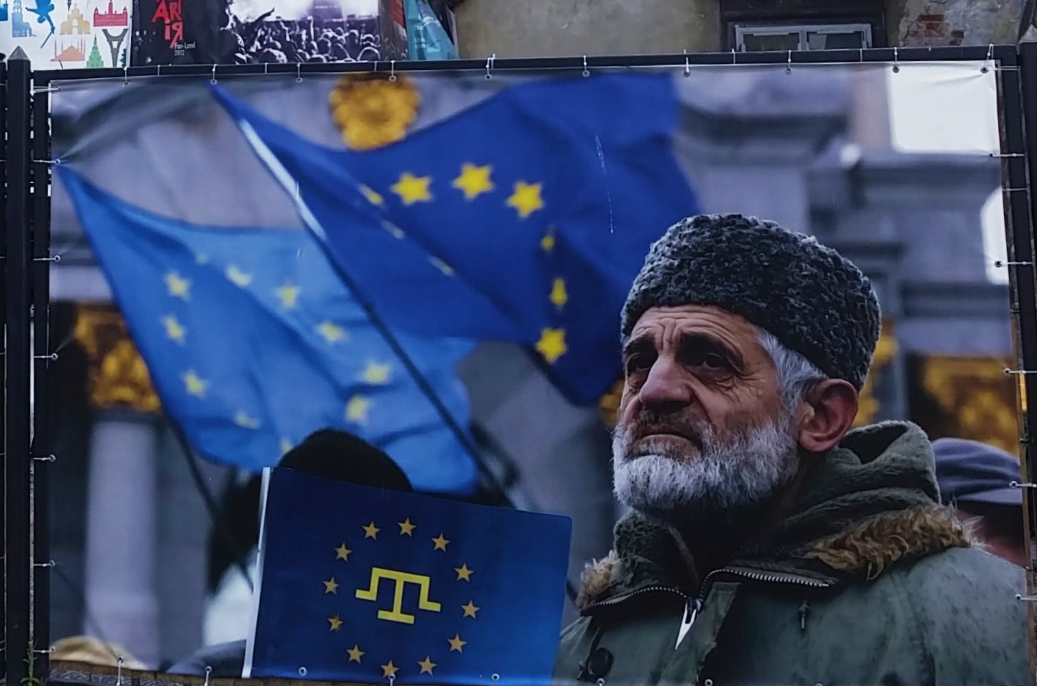 A large outdoor banner displays several blue flags with yellow stars resembling the European Union flag, waving in the background in front of a stone building. In the foreground, a person wearing a thick, fur‑trimmed winter coat and a textured hat holds a dark blue sign or flag featuring a yellow emblem that combines a stylized trident‑like symbol with a circle of stars. The banner is attached to a metal fence with cords, and the scene appears to be part of a public display or event.
