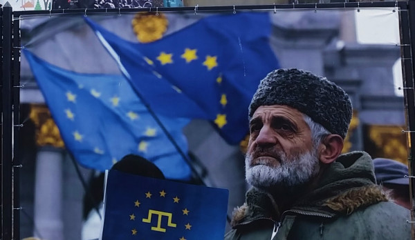 A large outdoor banner displays several blue flags with yellow stars resembling the European Union flag, waving in the background in front of a stone building. In the foreground, a person wearing a thick, fur‑trimmed winter coat and a textured hat holds a dark blue sign or flag featuring a yellow emblem that combines a stylized trident‑like symbol with a circle of stars. The banner is attached to a metal fence with cords, and the scene appears to be part of a public display or event.