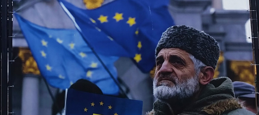 A large outdoor banner displays several blue flags with yellow stars resembling the European Union flag, waving in the background in front of a stone building. In the foreground, a person wearing a thick, fur‑trimmed winter coat and a textured hat holds a dark blue sign or flag featuring a yellow emblem that combines a stylized trident‑like symbol with a circle of stars. The banner is attached to a metal fence with cords, and the scene appears to be part of a public display or event.